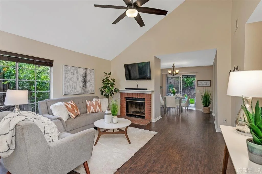 Living room with beige walls, a gray sofa with patterned pillows, a white armchair, a white rug, a brick fireplace with a mounted TV, plants, and a large window showing greenery outside. Adjacent dining area with a table, chairs, and chandelier.
