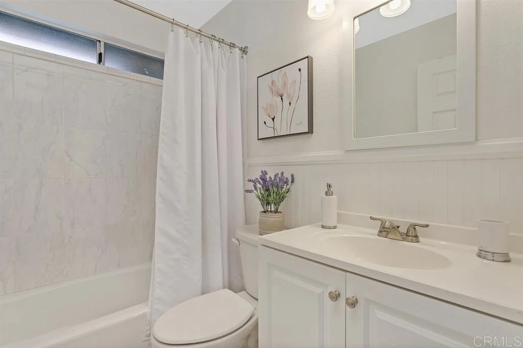 Small white bathroom with a bathtub, shower curtain, toilet, and vanity with a mirror, lavender flowers, and wall art