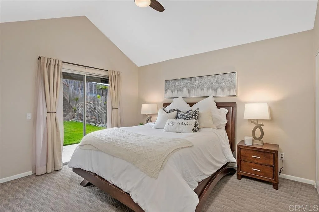 A cozy bedroom with a large bed with white linens and decorative pillows. There are two nightstands with lamps on either side of the bed. A sliding glass door with beige curtains leads to a backyard with green grass and a wooden fence.
