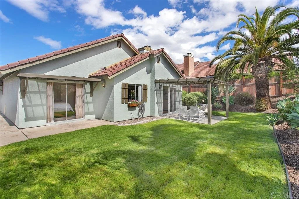 Backyard with green lawn, palm tree, patio area with outdoor dining table and chairs, house with gray exterior walls and red tile roof, blue sky with clouds.