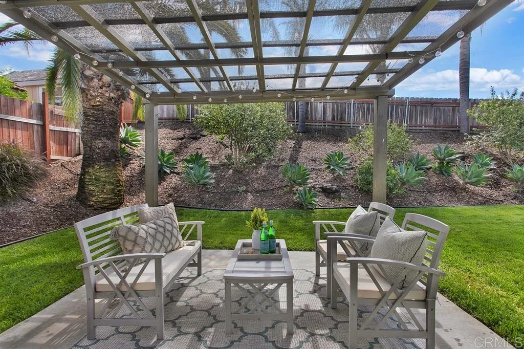 Outdoor backyard seating area with four white chairs, a matching table, and cushions, under a covered pergola with string lights, surrounded by a landscaped garden with succulents, palm trees, and a wooden fence.