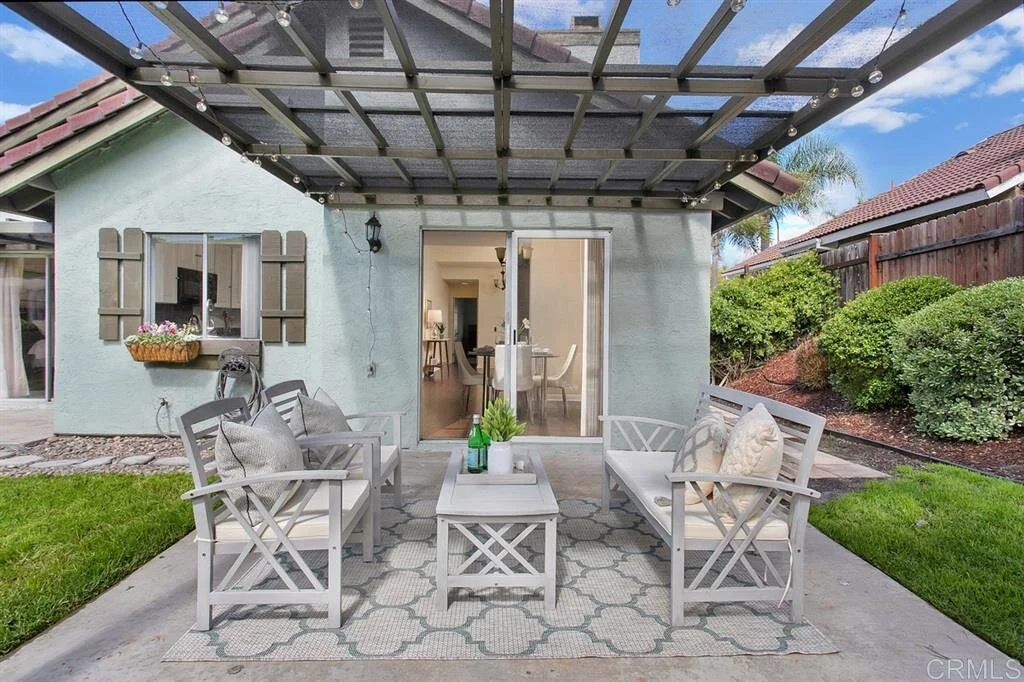 Outdoor patio with four white chairs, a matching coffee table, and cushions, under a transparent roof, with a house behind and a garden on the right.