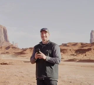 A man standing in a desert landscape with rock formations in the background, holding a smartphone and smiling.