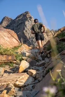 Hiker ascending a rocky mountain trail with large rocks and boulders, under a clear blue sky.