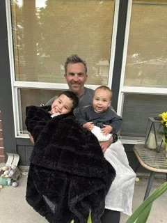 A man sitting on a porch with two children, holding a blanket, with a glass door and window behind them, a potted plant with yellow flowers on a small table, and some gardening tools on the ground.