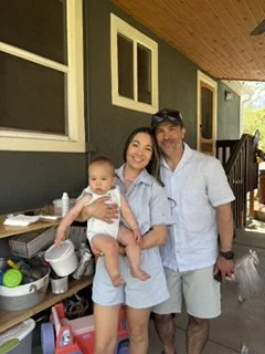 Three people: a woman holding a baby girl, and a man standing next to them outside a house with green walls and multiple windows.