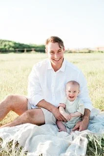 A man and a young child sitting together outdoors on a blanket in a grassy field, smiling at the camera.