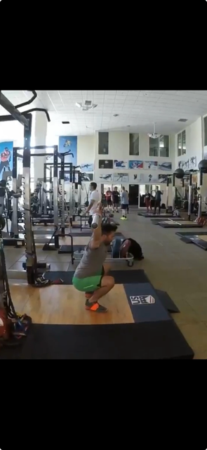A woman in athletic wear performing a squat exercise with a barbell in a gym, with others working out in the background.