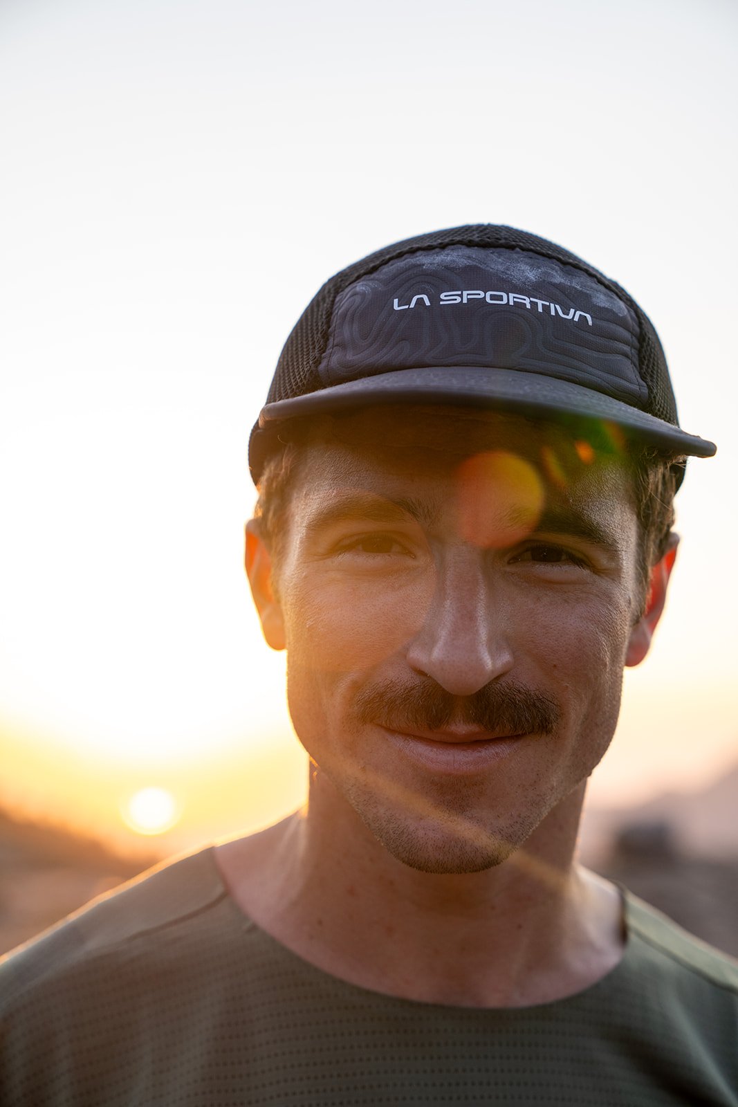 A young man with a mustache and light skin wearing a cap that says La Sportiva, smiling at the camera during sunset.