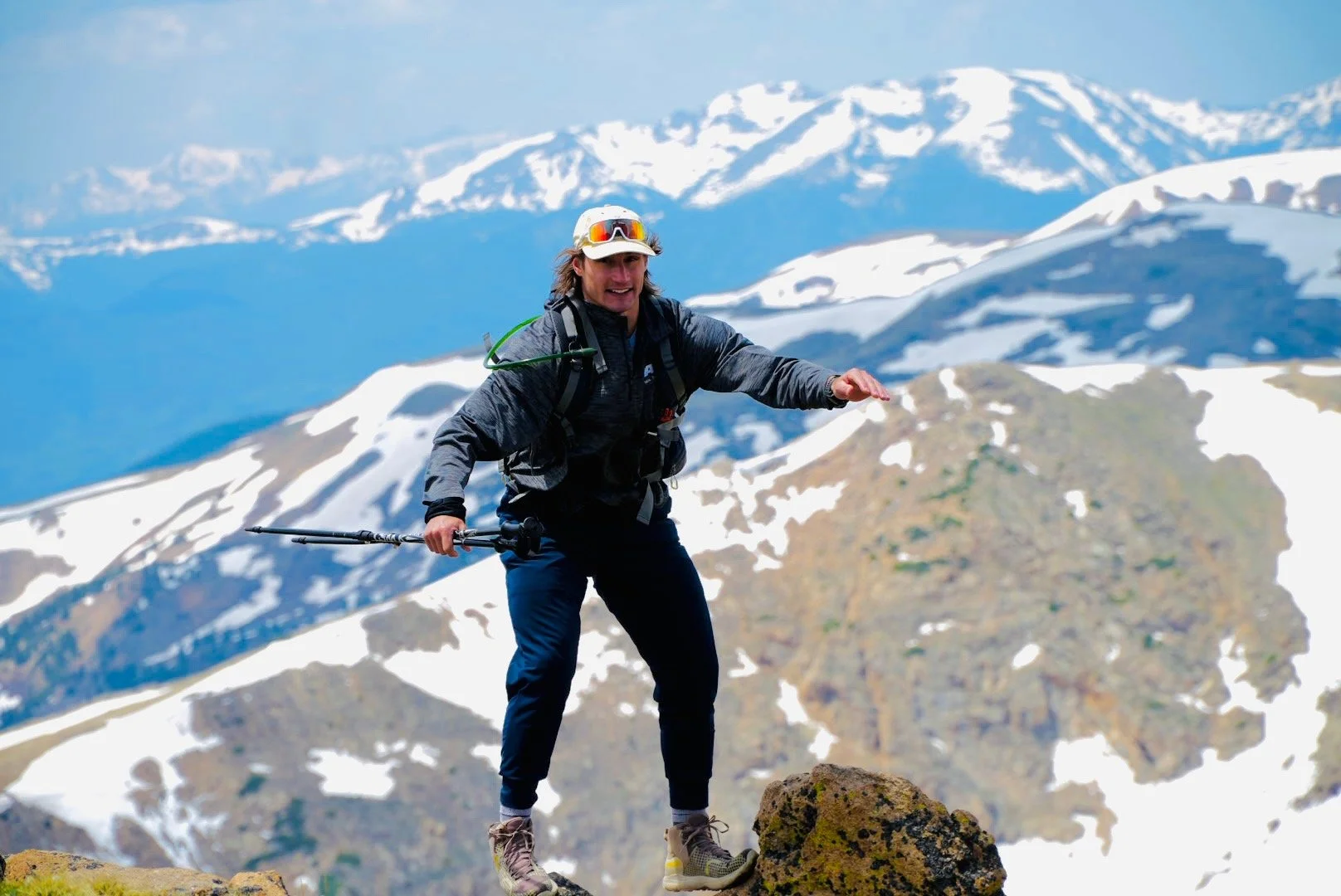 A man with long hair, wearing a hat, sunglasses, and outdoor gear, stands on a rocky mountain with snow-capped peaks in the background, holding a trekking pole and gesturing with his hand.