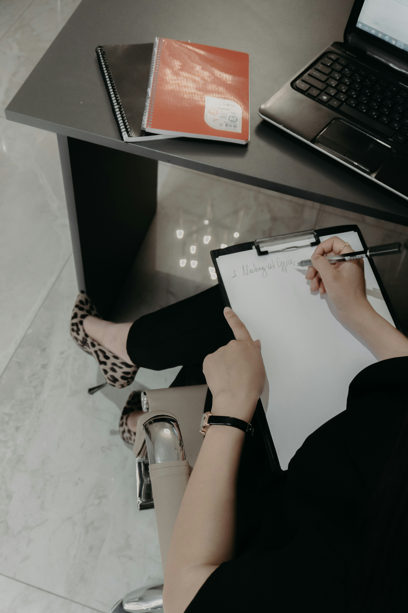 A woman sits at a desk writing notes on a clipboard while working through paperwork in an office setting.