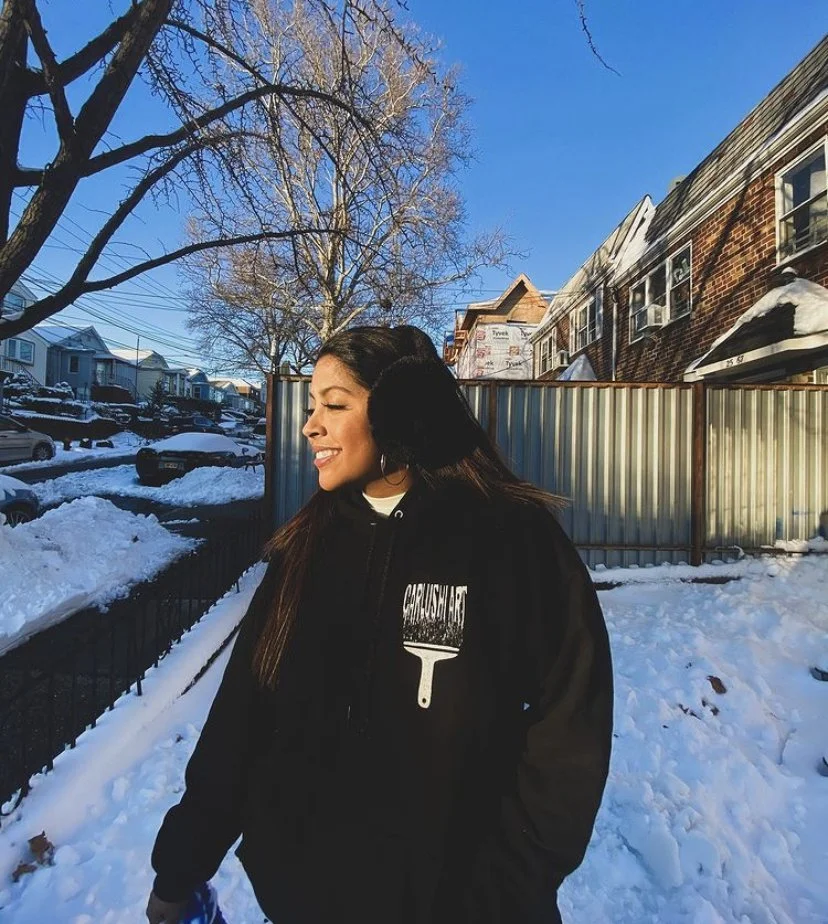 A woman with long dark hair smiling outdoors in a snowy neighborhood, wearing a black jacket with a paintbrush graphic and text, with houses, trees, and parked cars in the background.