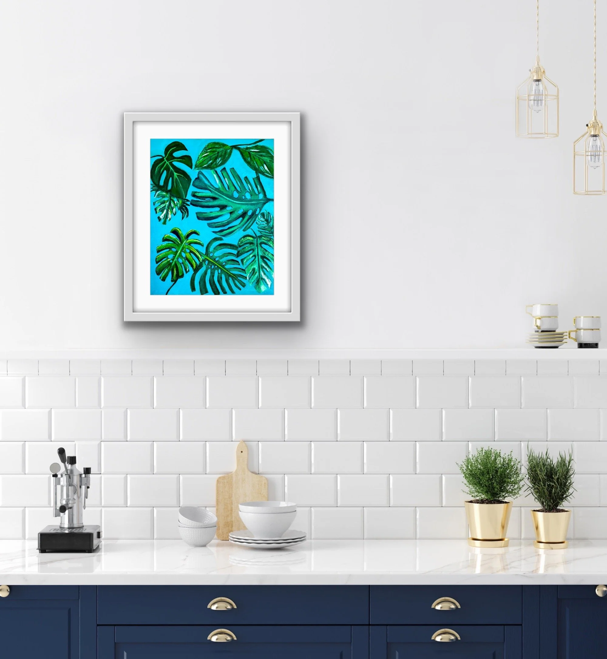 Contemporary kitchen with white subway tile backsplash, navy blue cabinets with gold handles, and a white marble countertop. A framed artwork of tropical leaves hangs on the white wall. Decor includes two small potted plants in gold pots, white dishes, and a cutting board, with hanging lights and some kitchenware.