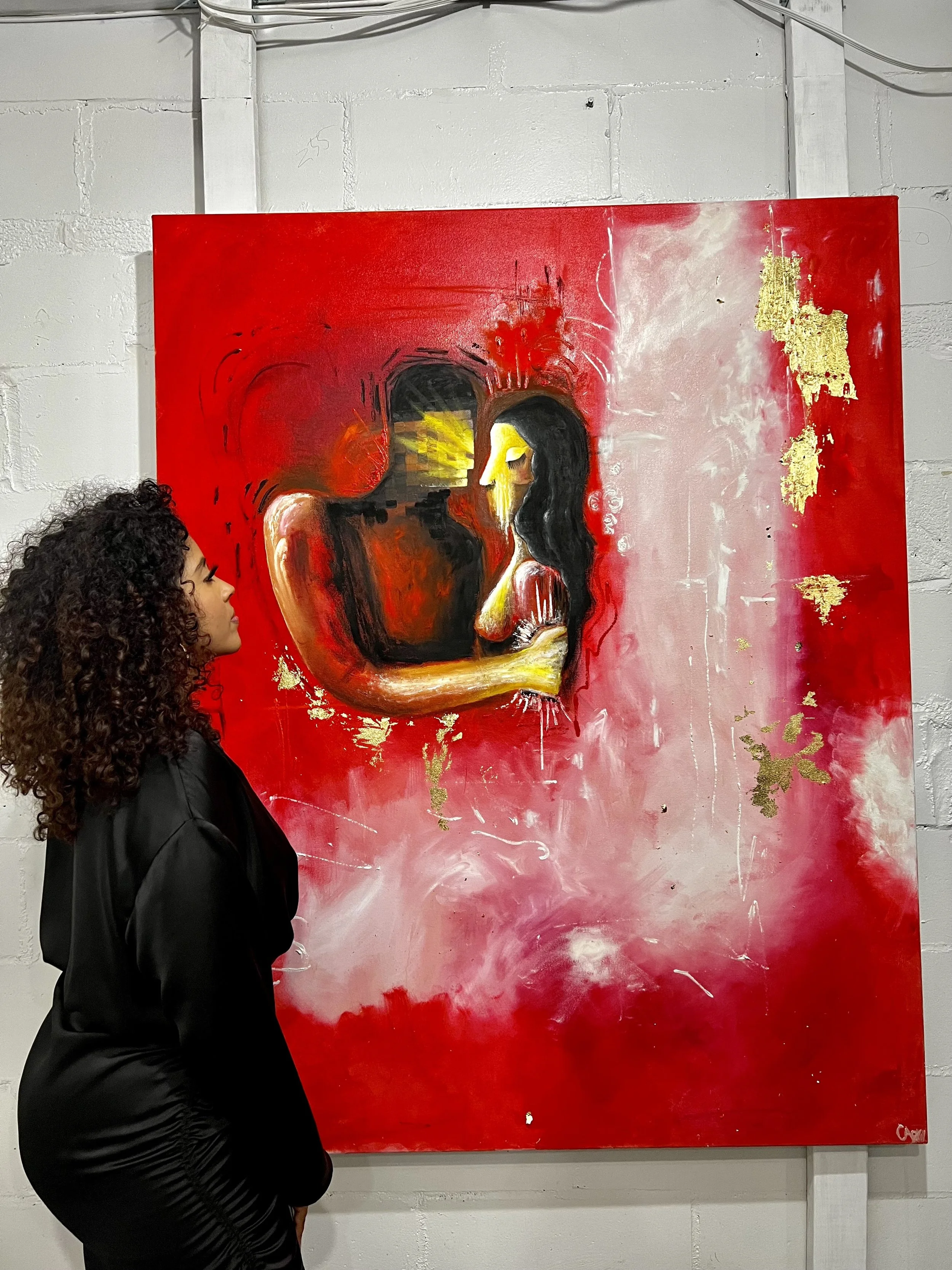 A woman with curly hair wearing a black jacket looking at an abstract painting of a couple embracing, with red, black, yellow, and white colors and gold accents on a large canvas in an art gallery.