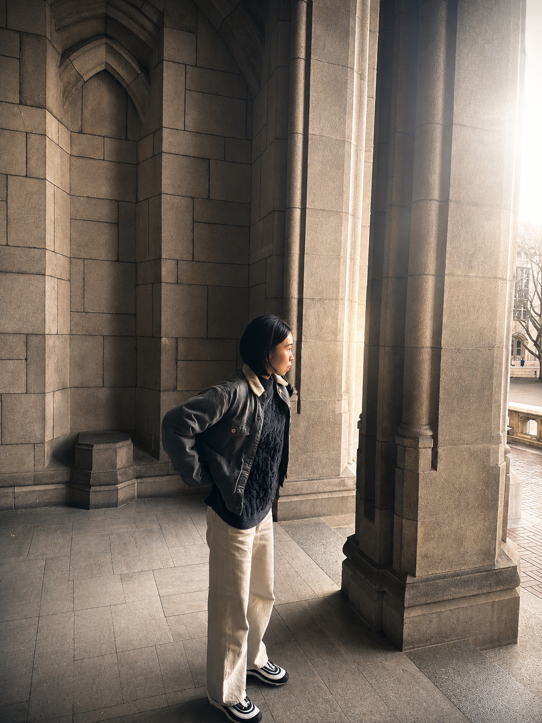 A female contemporary artist stands inside an old stone building with large columns, looking out towards a sunlit exterior with brick pavement and trees.
