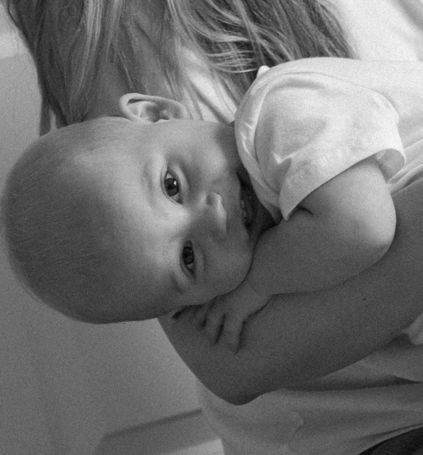 A baby with a shaved head resting on an adult's shoulder, looking at the camera with a curious expression.