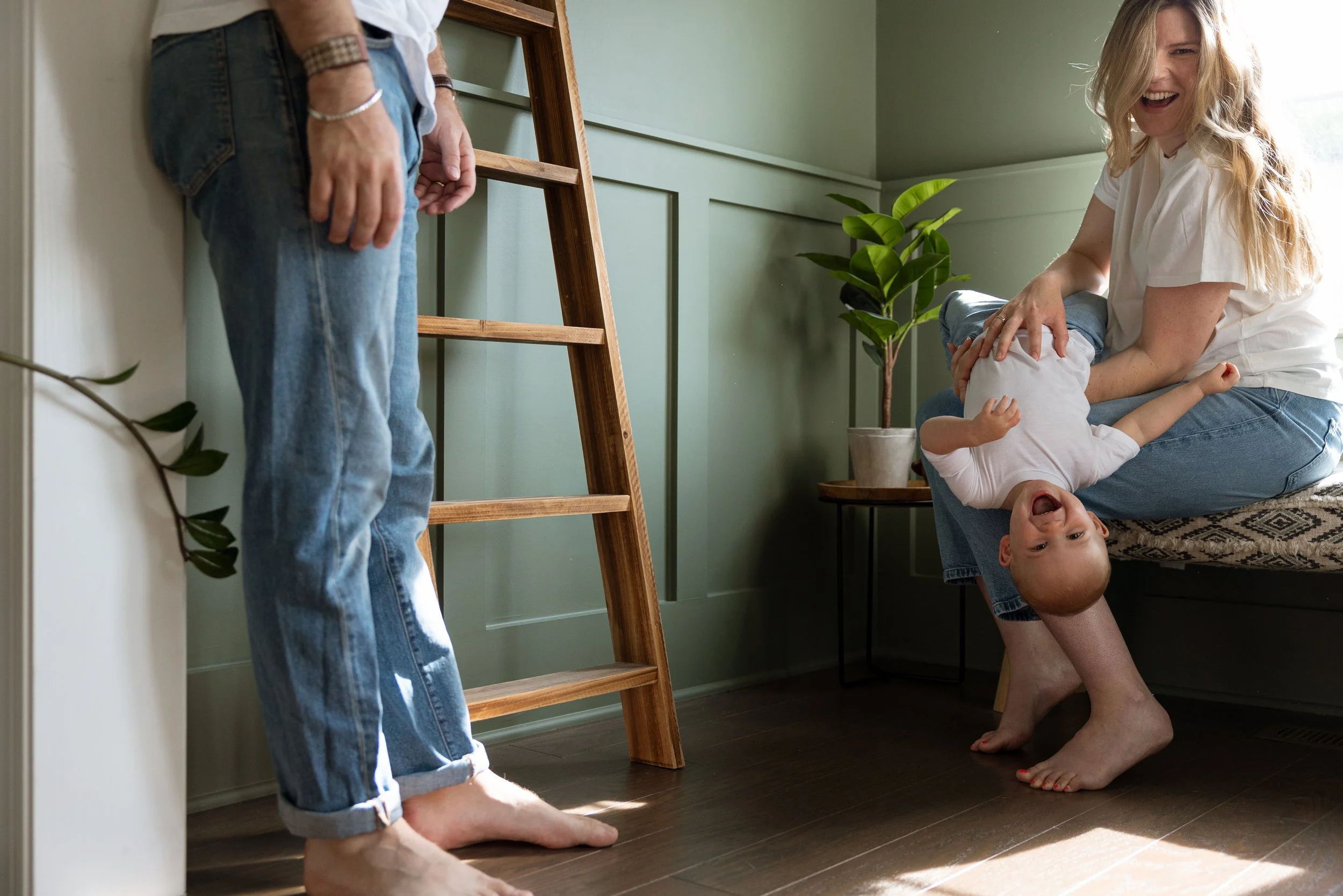 Woman playing with a young child who is hanging upside down in a room with green walls, a potted plant, and a wooden ladder.