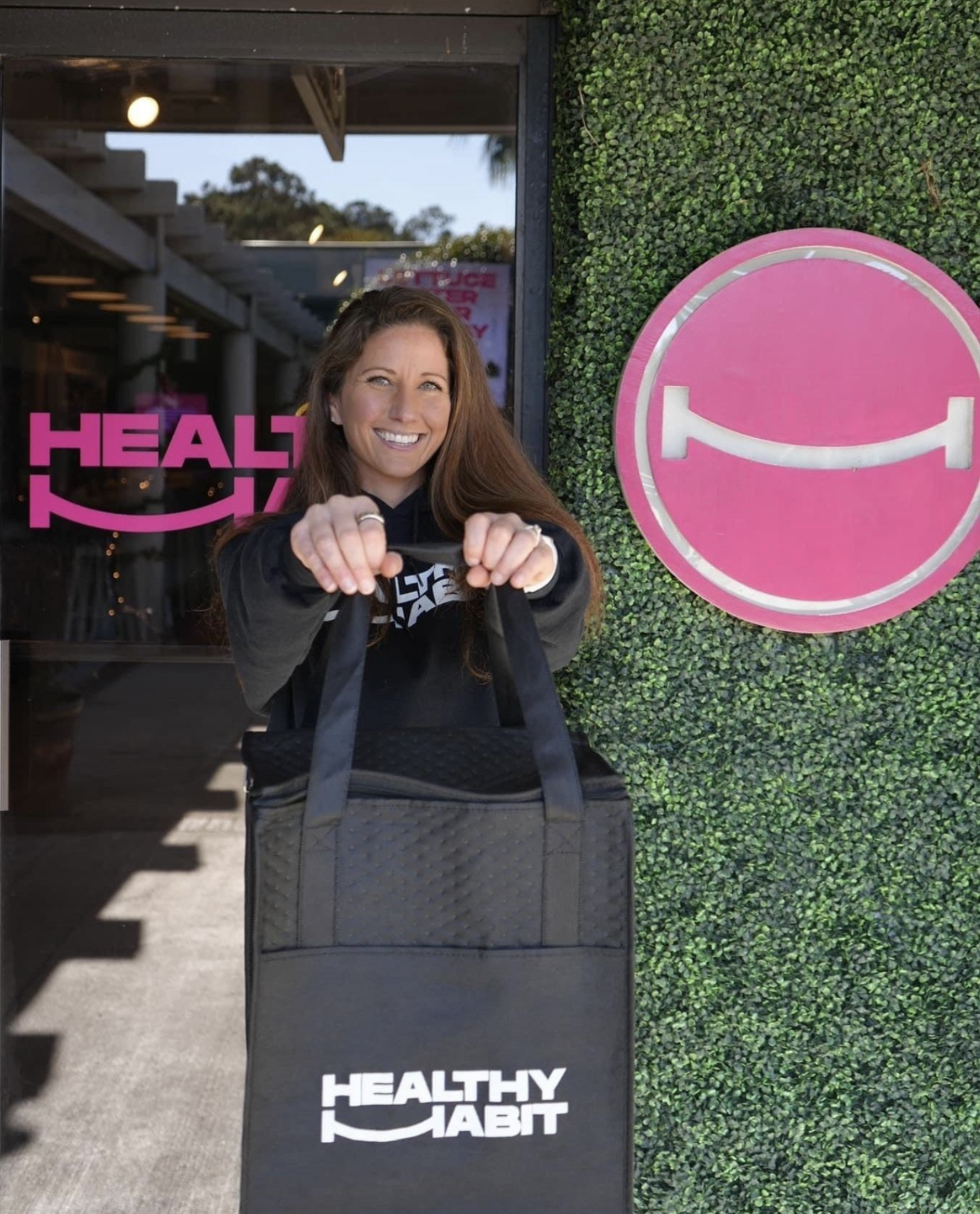 A smiling woman with long brown hair holding a bag in front of a storefront with a sign that says 'Healthy Habit' and a pink smiley face logo on a green hedge wall.