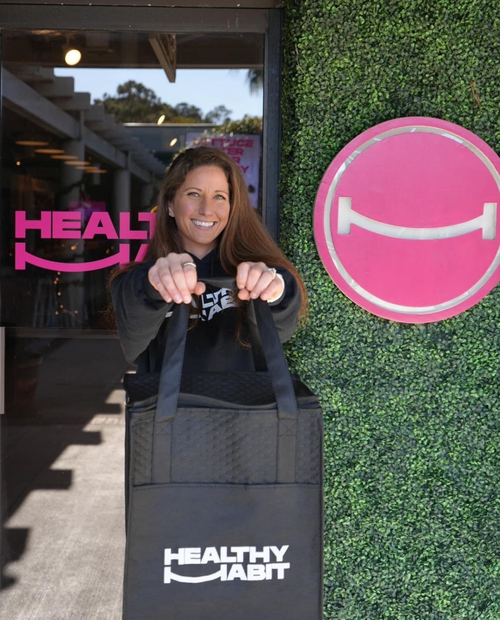 A woman with long brown hair smiling and pointing at camera while holding a black bag labeled 'Healthy Habit' in front of a green hedge wall and pink smiley face logo.
