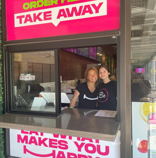 Two women working at a takeout restaurant counter, smiling, with a bright pink sign above that says 'ORDER HERE TAKE AWAY' and another sign below that says 'EAT MAKES YOU HAPPY.'