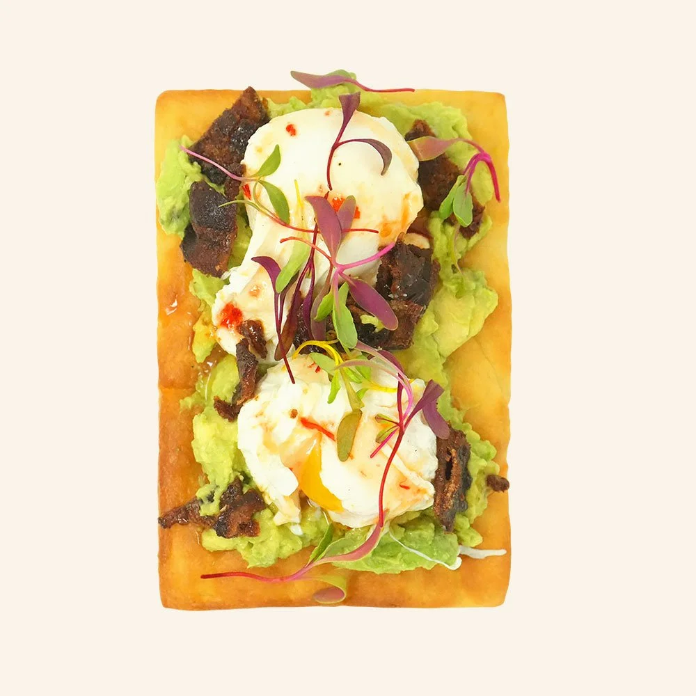 LOADED AVOCADO TOAST - Mashed avocado on house made organic focaccia bread, with candied bacon, two poached eggs, Calabrian drizzle and topped with local micro greens.