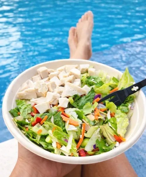 Person relaxing by a pool with legs visible, holding a bowl of salad with chopped chicken, lettuce, carrots, and other vegetables.