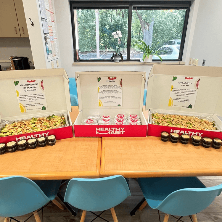 Three lunch boxes labeled 'Blackened Chicken Caesar,' 'Wraps,' and 'Dynamic Salad' with small jars of dressing in front, set on a table with blue chairs, near a window with potted plants outside.