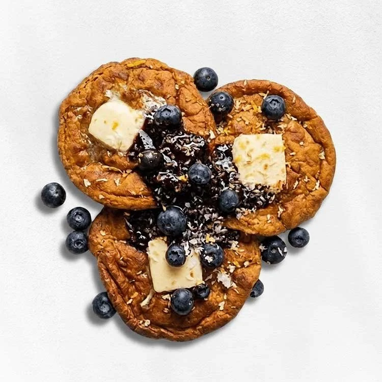 Three chocolate chip cookies topped with blueberries, chocolate syrup, and white chocolate pieces on a white background.