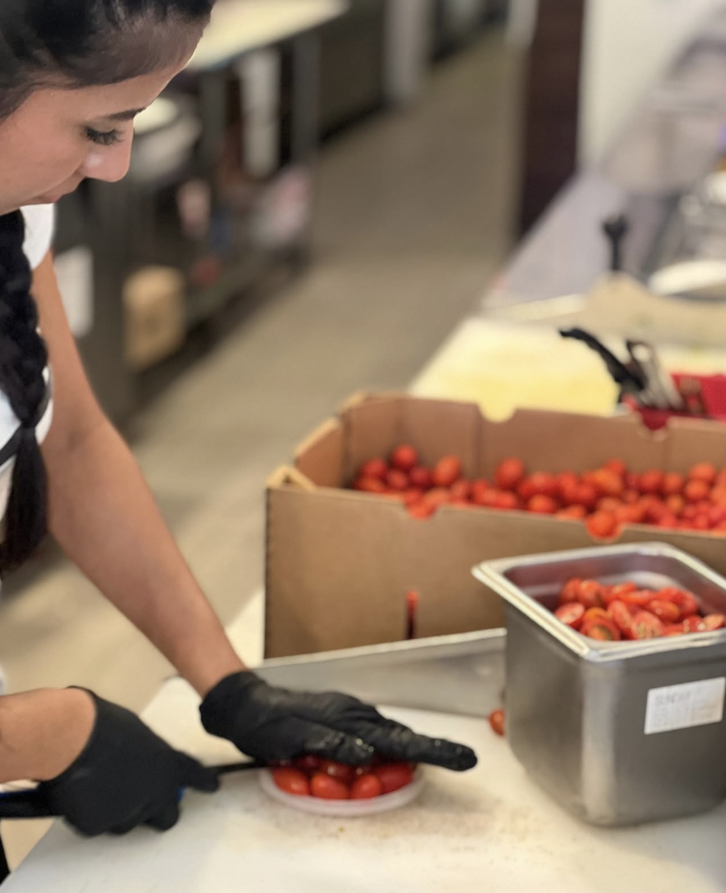 Close-up of a person wearing black gloves, handling red cherry tomatoes on a white plate in a kitchen or food preparation area. There are boxes of tomatoes and other kitchen items in the background.