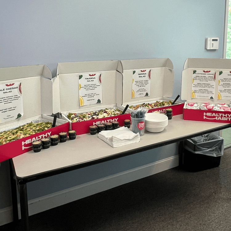 A table set up with three open salad boxes labeled "Healthy Habit" containing different salads, along with small dressing cups, white bowls, napkins, and utensils, in a room with a light blue wall and a thermometer on the wall.