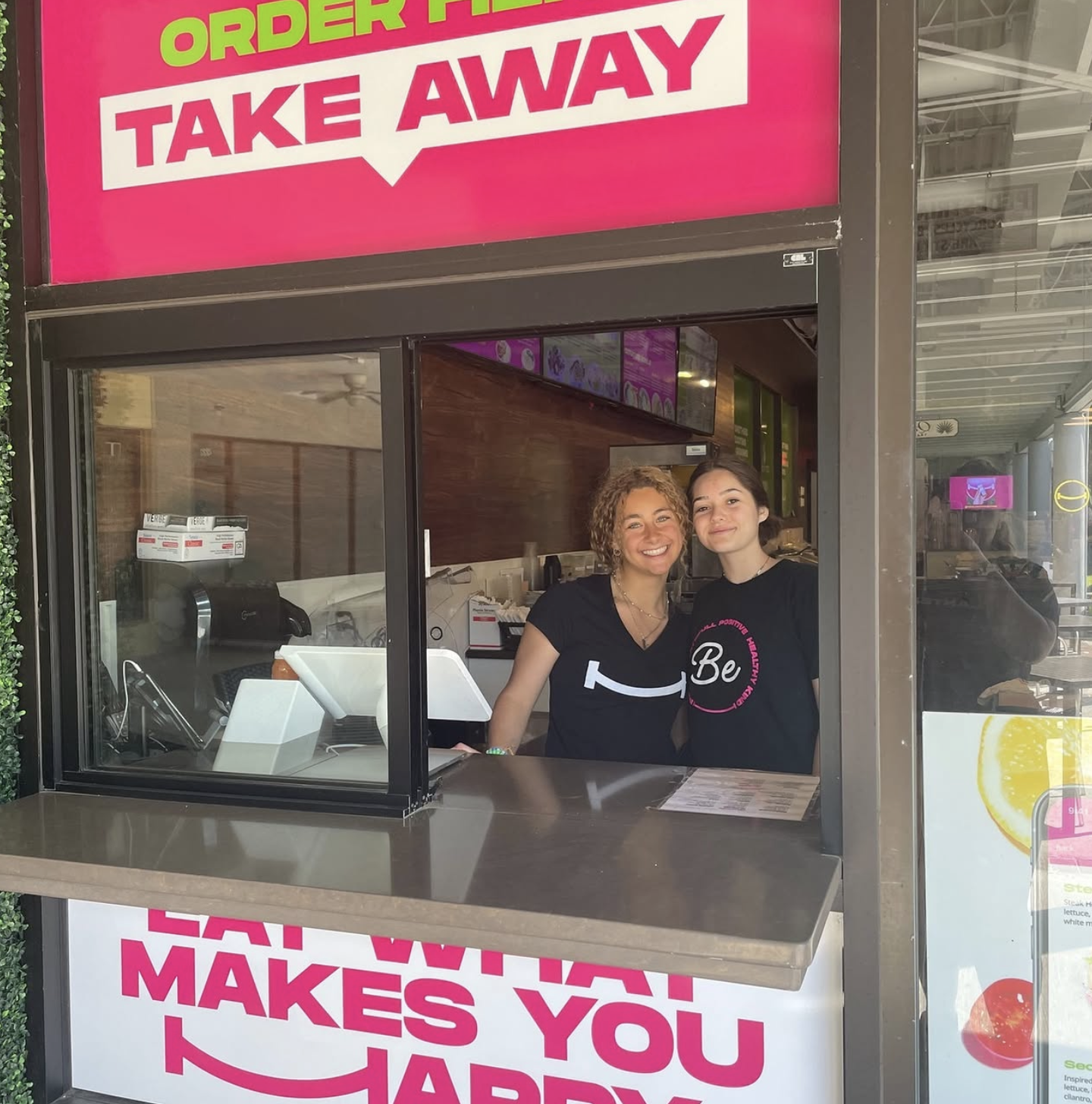 Two women smiling inside a colorful takeout food stand, with bright pink and green signage that says, 'Order Here, Take Away.'