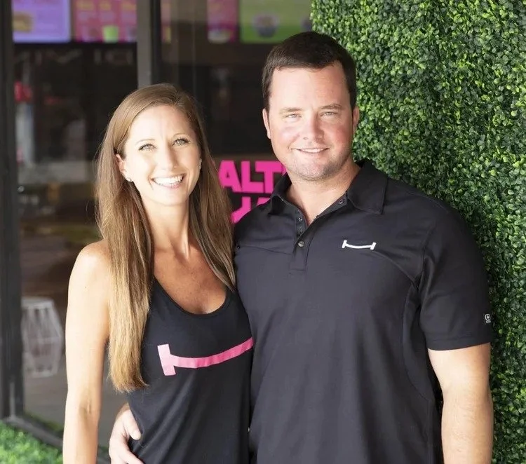 A smiling woman with long brown hair wearing a black sleeveless top, standing next to a smiling man with short dark hair wearing a black polo shirt, near a green hedge outdoors.