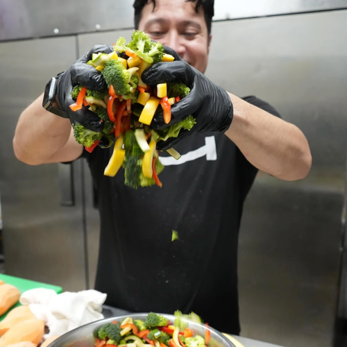 Man wearing black gloves holding a handful of fresh mixed vegetables, including broccoli, yellow bell peppers, and carrots, over a kitchen counter.