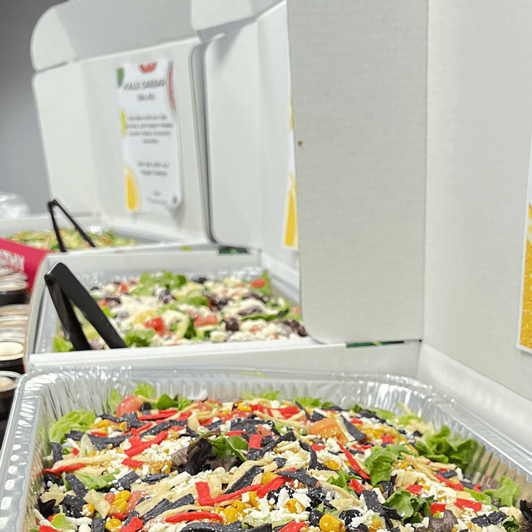 Tray of freshly made salad with lettuce, corn, black olives, shredded cheese, and sliced red peppers at a buffet.
