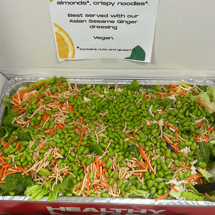 Fresh vegan salad with lettuce, shredded carrots, edamame beans, and shredded vegetables in a tray, with a sign in the background promoting almonds and crispy noodles with Asian sesame ginger dressing.