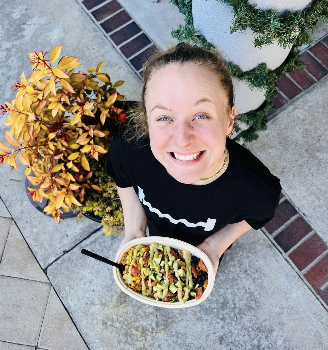 A young woman with blonde hair and blue eyes smiling joyfully, holding a bowl of salad topped with cucumbers, cherry tomatoes, and cheese, standing outdoors on a concrete patio next to a potted plant with yellow and red leaves, and a white planter wi