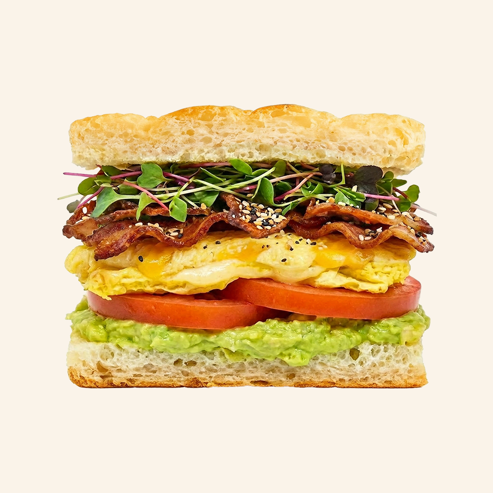 BREAKFAST SANDWICH - Mashed avocado topped with a cheddar jack cheese omelette, crispy bacon, tomato, everything bagel seasoning and local microgreens on organic focaccia. 