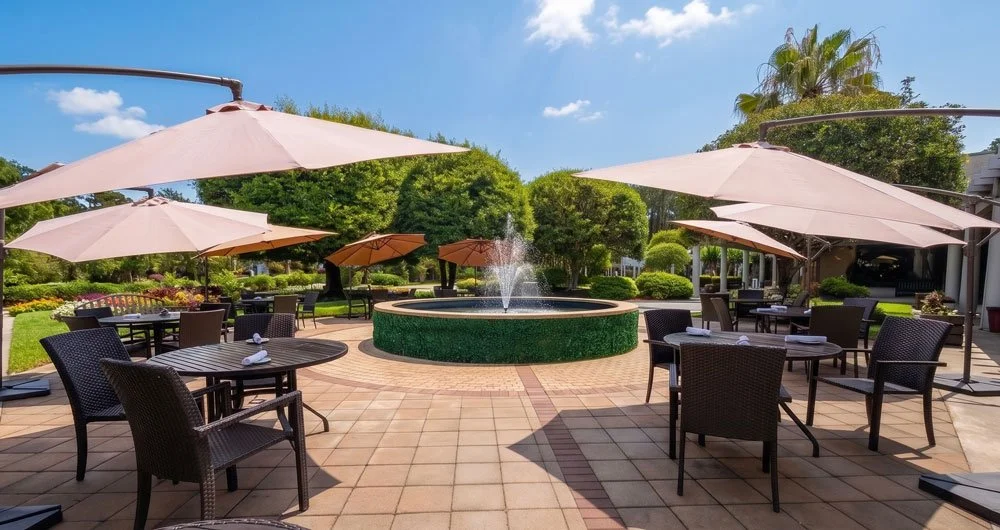 Outdoor restaurant patio with tables, chairs, umbrellas, a fountain, and lush greenery under a partly cloudy sky.