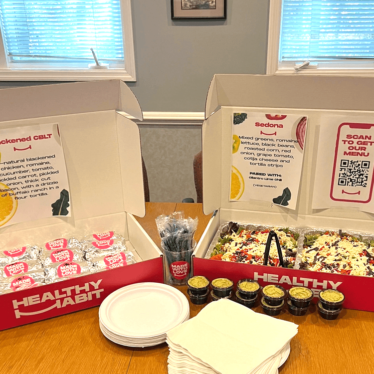Pizza boxes with salads, foil-wrapped items, small containers of dressing, paper napkins, and plates on a wooden table.