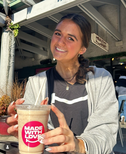 A smiling young woman holding an iced coffee in an outdoor cafe setting.
