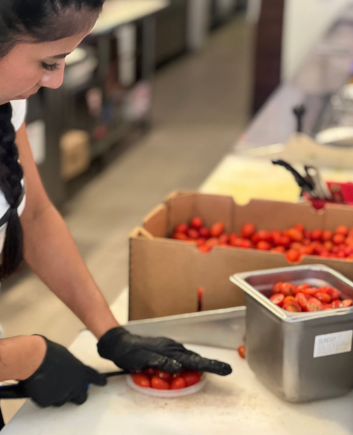 A person wearing black gloves slices tomatoes in a kitchen or food preparation area with boxes of tomatoes nearby.