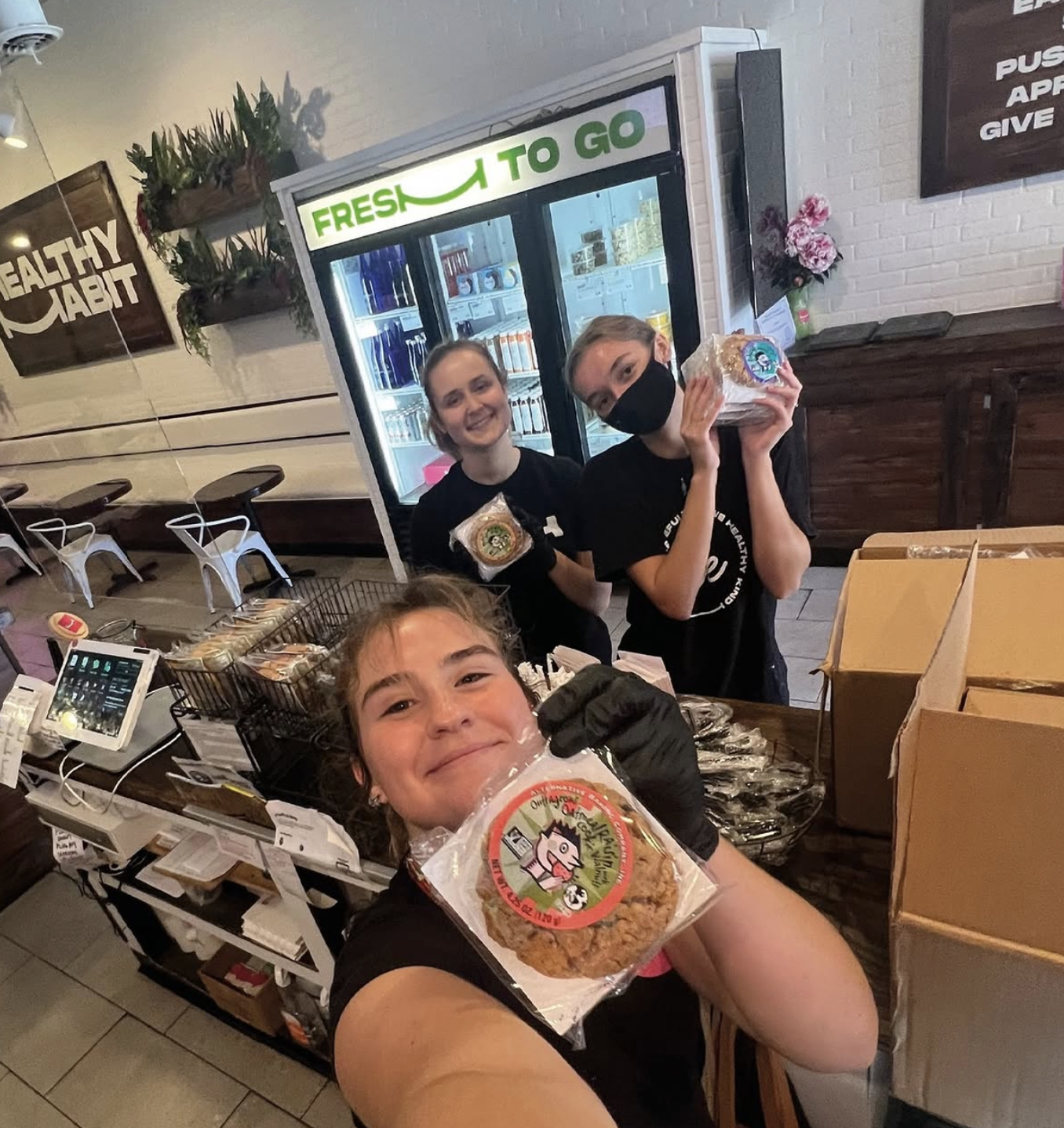Three smiling women working at a bakery counter with a display of bakery products, holding packaged cookies, with a beverage fridge and signs on the wall in the background.
