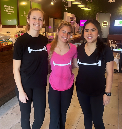 Three smiling women in black and pink promotional shirts standing inside a cafe or restaurant.