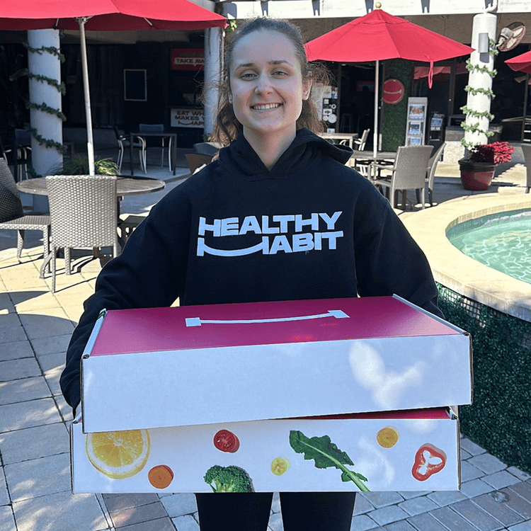 A young woman with a big smile holding a box, standing outdoors near a pool with tables and red umbrellas.