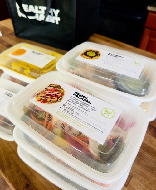 Plastic meal containers on a wooden surface, labeled with dishes such as Greek falafel, with a green tote bag in the background.
