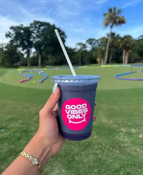 A hand holding a purple smoothie in a clear plastic cup with a white straw, outdoors on a sunny day, with colorful tubular obstacles on a grassy area in the background.