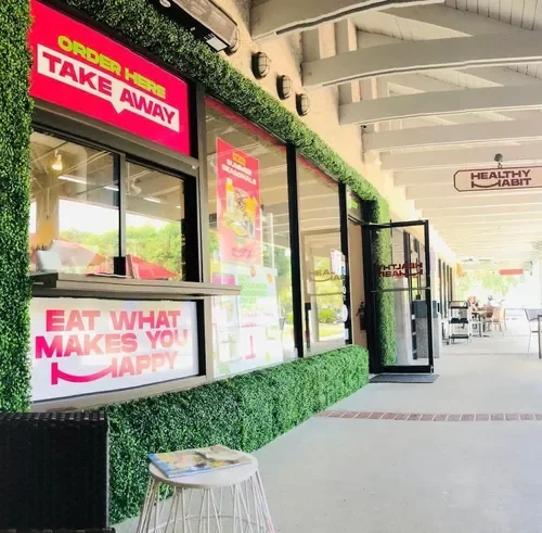 A storefront with a pink sign reading 'Order Here Takeaway' and a window display with a message 'Eat what makes you happy'. Outside the store, there is a small table with magazines and a few chairs in the covered shopping center.