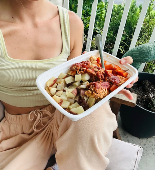 A woman in a yellow tank top and beige sweatpants holding a white tray with food, sitting outside near a black pot and green plants.