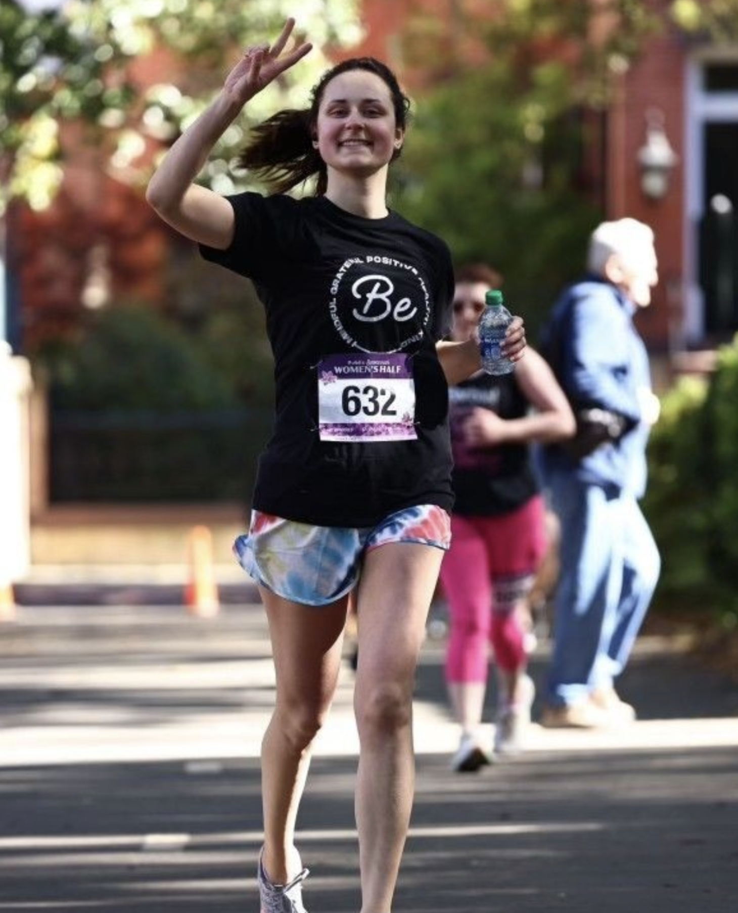 Young woman smiling and making a peace sign while running in a race, holding a water bottle, with other runners and trees in the background.