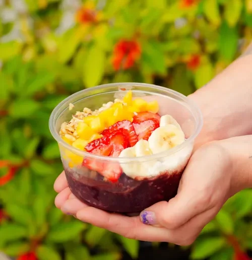 A hand holding a clear plastic cup filled with a layered dessert topped with strawberries, diced mango, banana slices, and granola, with a blurred green plant background.